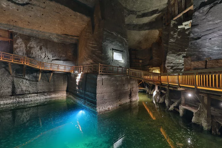 Ein Blick auf den wunderschön beleuchteten unterirdischen Salzsee in der Erazm Barącz-Kammer. Im Hintergrund sind die Salzsäule und die dicken Salzschichten an den Wänden zu sehen. Um den See herum führt ein Holzsteg für Touristen.