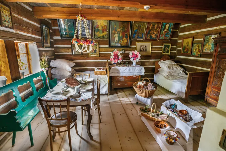 A typically furnished interior of one of the wooden peasant cottages in the open-air museum. It contains wooden chairs, a table, beds with piles of pillows, numerous sacred paintings hanging on the walls made of wooden beams.