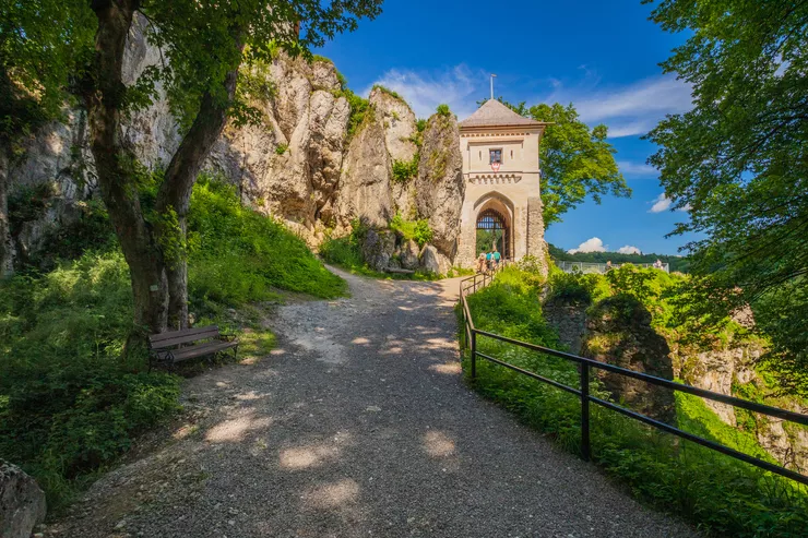 Blick auf den Turm mit dem Haupteingangstor, der von Kalksteinfelsen umgeben ist und zu den Ruinen der Burg Ojców führt. In der Ferne sind Touristen zu sehen, die die Burg verlassen. Vor dem Tor sieht man eine Straße mit Bäumen und Gras an den Seiten. Der Himmel ist blau.