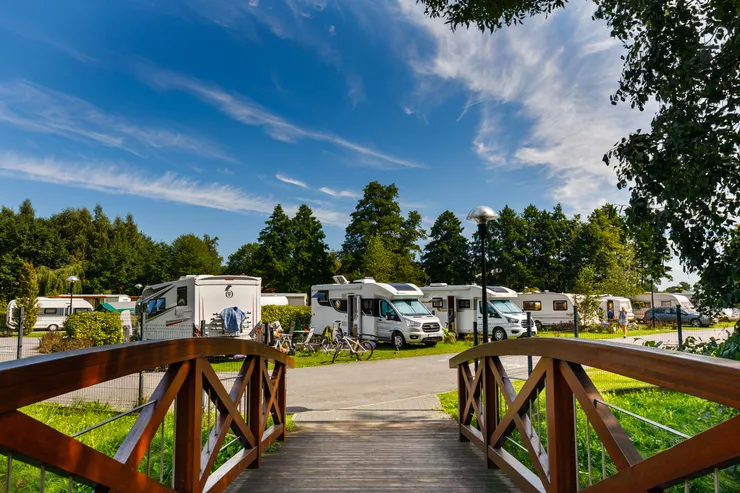 A view over a wooden bridge with wooden railings into a camper van park on the other side of a road. There are several white camper vans parked in front of a row of tall trees. The summer sky is blue with some long white clouds.