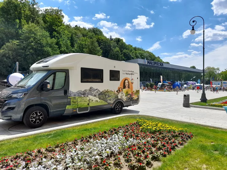 A camper branded with Małopolska commercials is standing in the main promenade of Krynica, behind a flower patch. Visible behind it is the main pump room with deckchairs standing in front of it among walking people. The blue sky is speckled with small clouds.