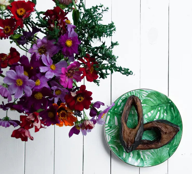 Two slices of roasted carp lie on a green plate on a table made of rough white planks. Standing to the left above it is a bouquet of colourful flowers with several boxwood twigs.