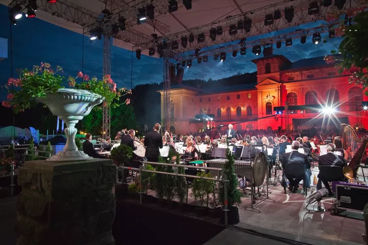An orchestra are playing on an artificially lit open-air stage. The background is taken by the Old Spa House of Krynica backlit by the invisible moon on very dark blue sky.