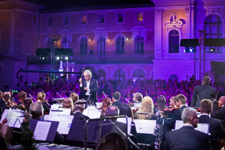 Blick auf die Bühne, auf der das Orchester ein abendliches Konzert spielt. Im Hintergrund ist das blau beleuchtete Gebäude des Alten Kurhauses zu sehen. Die gesamte Bühne mit den Musikern ist ebenfalls beleuchtet.
