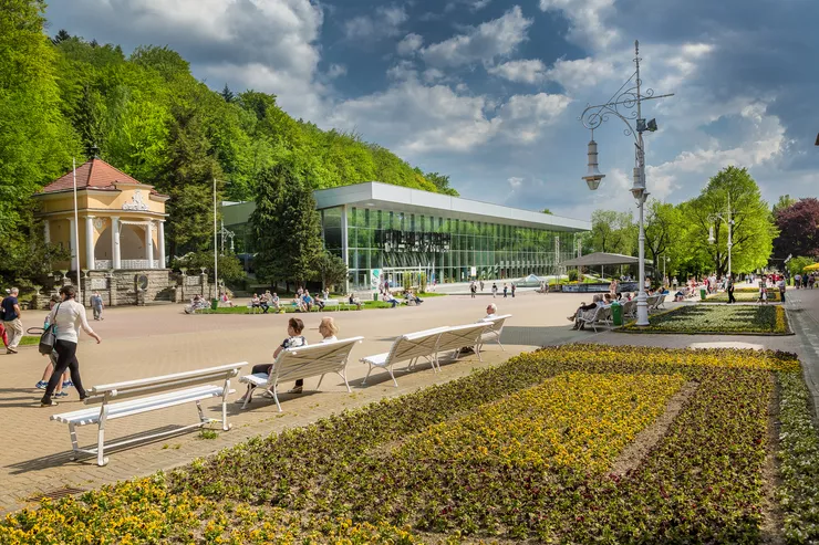 A view of a historical bandshell and the New Pump House on the far side of Krynica Spa Promenade. Some tourists are walking along the wide sunlit promenade, while others sit on white benches. The foreground is taken by a carpet of flowers.