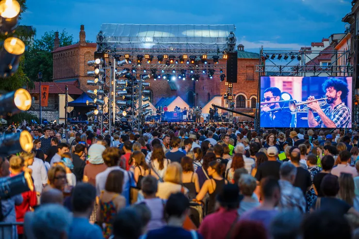 A band are performing on a large open-air stage in the centre of the picture. Two musicians playing trumpets can be seen on a bright TV wall to its right. In the square in front of the stage, hundreds of people are listening to the evening concert, there is a large historical brick building behind the stage.