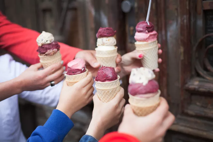 A view of six different hands holding cones with two and three scoops of colourful ice cream against of a dark wooden door in the background.