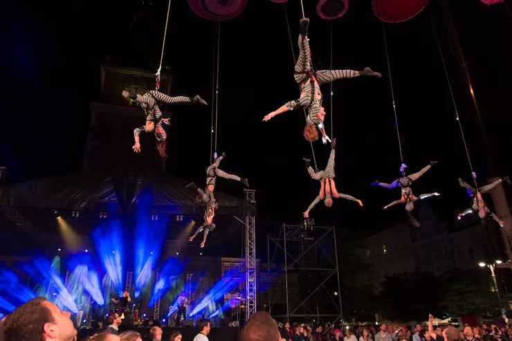 It’s a dark night, spotlights highlight six actors hanging head-down from the ropes high over the crowd. A stage lit with blue beams can be seen in the bottom left corner.