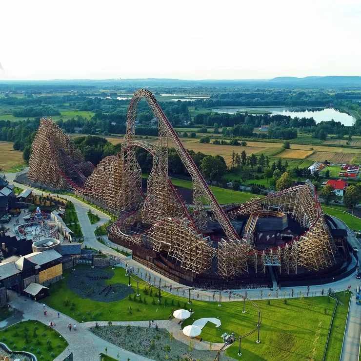 A view of the world’s tallest wooden roller coaster built of wood and metal only. Cars with people are starting the slide from its tallest point. A section of the amusement park with some other attractions is in the foreground, while the distant plane shows pleasant countryside with a lake and a town in the distance.