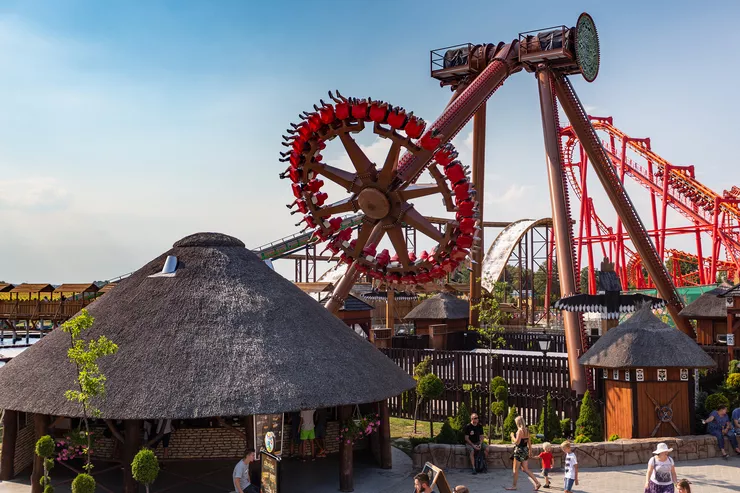 A view of a massive spinning swing, with a wheel of almost 27-metre diameter. People are seated in the wheel. Below, in the foreground, children with their parents are walking along the path among the amusement park’s huts. The sky is clear.