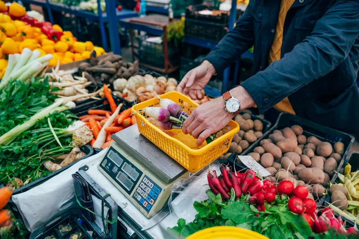 A low shot of a vegetable stall with bell peppers, parsley, carrots, onions, potatoes, and radishes with a centrally standing electric scale. A seller is placing a bunch of radishes in the orange plastic basket on top of it.