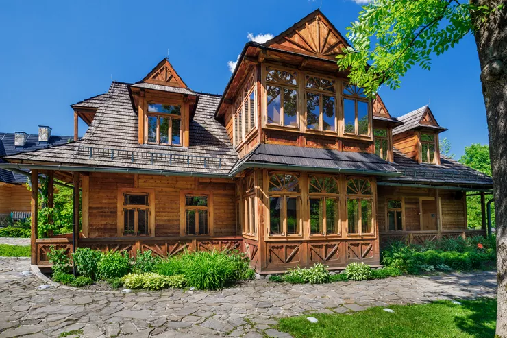 A view of a beautiful wooden building of the Karol Szymanowski Museum in Zakopane style, with tall rooves and sculpted decorations. There are some flowers and low shrubs growing by the walls of villa, separating it from a stone path in the foreground. The sky is blue and cloudless.