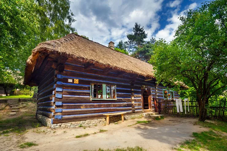 A view of a thatched wooden building standing in a regional open-air museum with construction features characteristic of the folk culture of Western Krakowiacy. It stands among leafy trees under a blue sky sprinkled with white clouds.
