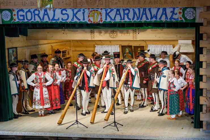 A large group of men and women dressed in traditional Highlander costumes are standing on a stage with wooden walls. Three men in the front are holding alpenhorns that they are playing. The banner above the stage reads “Highlander Carnival”.