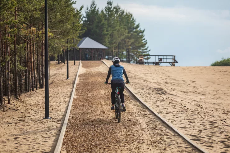 A view of a young woman riding a bicycle along a purpose-built path in the desert. She is wearing sportswear and a helmet. At the end of the path, there is a shelter and a metal platform. Some trees and lamp posts stand to the left of the path, and the sky is cloudy.