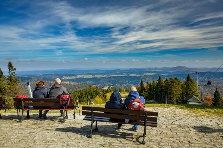 A view of four people sitting on wooden benches, facing away and admiring the beautiful landscape with many lines of wooded hills and mountains below. The cables and a gondola of the Jaworzyna Krynicka cable car are visible on the right side. The sky is covered with wisps of white clouds, and a high cloud bank closes the perspective, pretending to be distant mountain range.