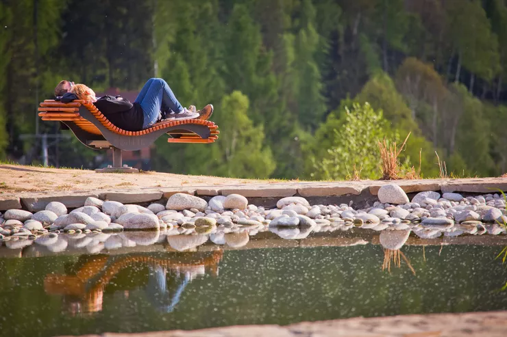 A view of two people relaxing among nature on a sophisticated sun lounger. They are mirrored in a small water reservoir with pebbles arranged along the edge taking the front of the photo, while the background consists of a wall of trees.