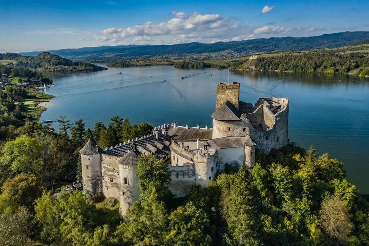 A drone-eye view of a limestone castle standing on a hill among plenty of trees. Below and behind it, there is a lake, on whose far bank the ruined Czorsztyn Castle stands. The sky above the reservoir and distant line of hills is blue with tufty clouds.