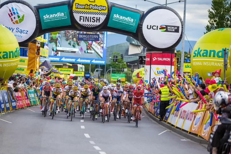 A view of a large group of cyclists passing through the starting gate as they start the race. There are many advertisements on the gate and on two oversized balloons standing behind clusters of spectators, who can be seen cheering on both sides of the track.