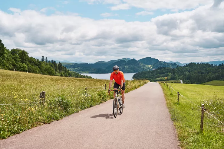 Blick auf einen Mann in Sportkleidung beim Radfahren. Entlang des Radwegs wächst auf beiden Seiten hohes Gras. Im Hintergrund sind der Czorsztyńskie-See und zahlreiche bewaldete Hügel zu sehen. Der Himmel ist blau mit vielen wallenden Wolken.
