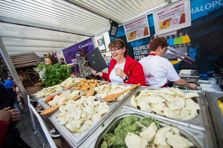 Two mature women operate the pierogi stand at the Małopolska Taste Festival. Displayed before them are metal containers with pierogi with various stuffing. There are wooden spoons lying on the pierogi. Advertising posters behind the women serve as the background, along with a roof made of translucent plastic panes above their heads.