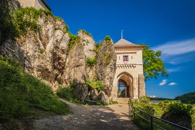 Blick auf den steinigen Weg, der zum Haupttor der Burg führt. Auf der rechten Seite des Weges befindet sich ein Metallgeländer. Das Eingangstor befindet sich dicht an den Felsen. Der Himmel ist blau.