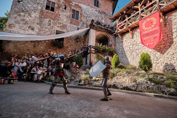 Auf dem Bild kämpfen zwei Ritter in Rüstungen gegeneinander. Um sie herum sind Teile der Burghofes zu sehen. Auf den Stufen, im Tor und auf dem hölzernen Balkon sind Menschen versammelt, die das Schauspiel beobachten.