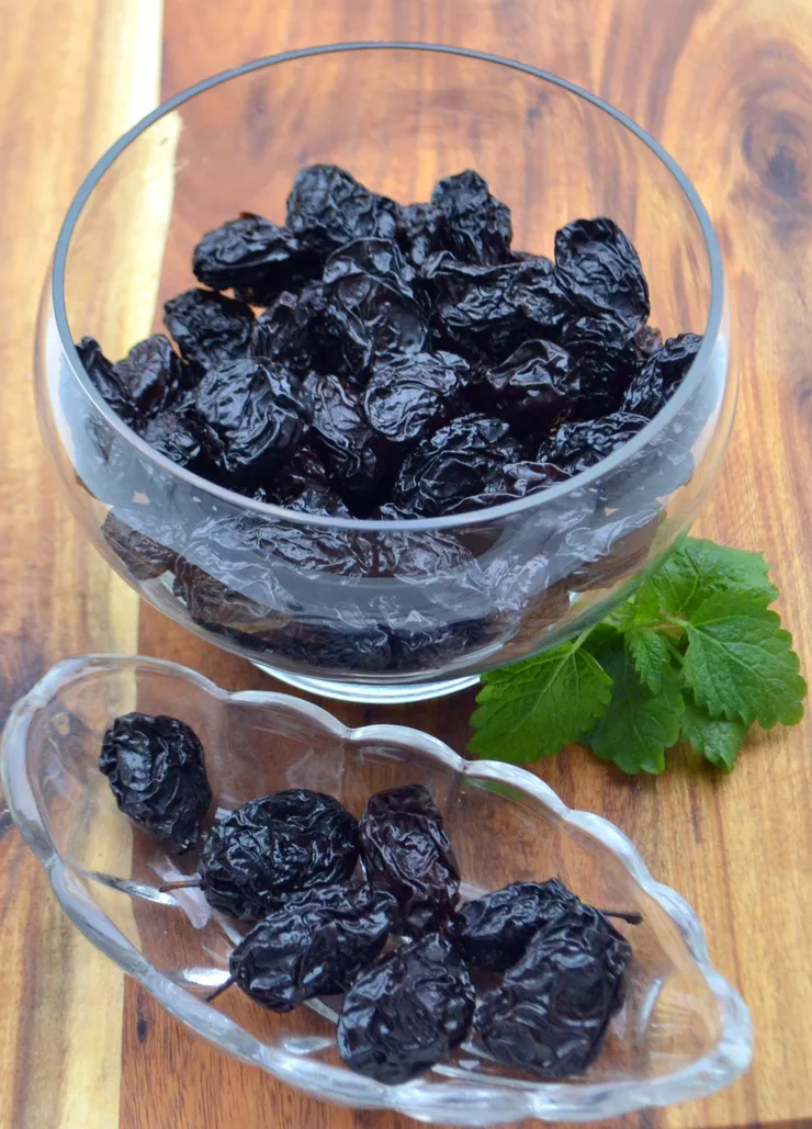 View of two glass bowls of suska sechlońska dried and smoked plums on a wooden plank. Lying beside the top round bowl are several mint leaves.