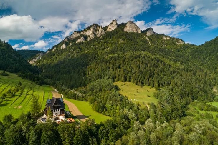 A drone-eye view of the sunlit PTTK Tourist Shelter Trzy Korony among the green fields and meadows leading up towards the edge of the forest, from over which the jutting rocks of Mt Trzy Korony are peering. There are plenty of white clouds in the sky.