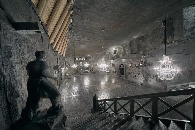 View of a huge salt chamber – chapel enclosed with a wall with an altar, whose walls bear numerous sculptures and bas-reliefs. Crystal salt chandeliers hang from the ceiling. There is a salt figure of a man, facing the chapel and standing with his back to the viewer, on the left-hand side on the first plane, above the stairs leading to the chapel.