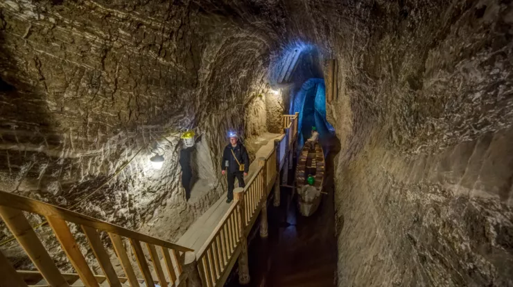 A miner walking along a wooden gallery with stairs along the left wall that curves up into a ceiling. Below him, there is a boat on an underground brine lake. The right-hand side is taken by a wall of rock or rock salt.