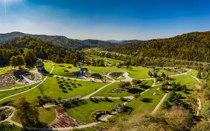 A drone-eye view of beautiful sensory gardens crisscrossed by many paths. They are surrounded by wooded hills under a clear blue sky.
