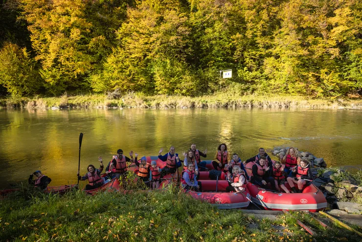 View of a river, with red rubber rafts and canoes filled with people wearing life jackets, about to start the rafting adventure, on its bank. The trees on the other bank are lit up by the sun.