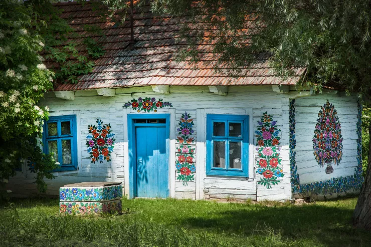 A white, wooden cottage with a tiled roof takes the whole picture. All its walls and the low well in front of it are decorated with colourful floral motifs. Its windows and door are blue. The whole is vignetted by tree branches and a grass lawn.