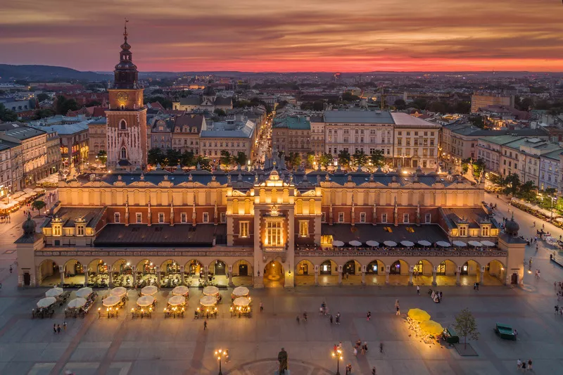 An aerial view of Kraków’s Main Market Square, with the brightly illuminated Cloth Hall taking the centre of the shot. Standing tall behind its left wing is the Town Hall Tower outlined against the evening orange-red sky. The foreground is speckled with a few rows of restaurant tables under umbrellas.