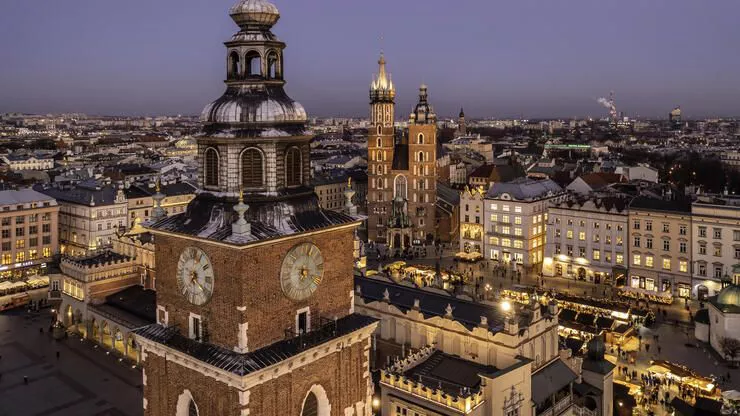 Eine Luftaufnahme des Krakauer Marktplatzes am Abend. Im Vordergrund ist der obere Teil des Rathausturms mit der Uhr zu sehen. Dahinter die Tuchhalle, die Marienkirche und die Altstadthäuser mit ihren beleuchteten Fenstern.