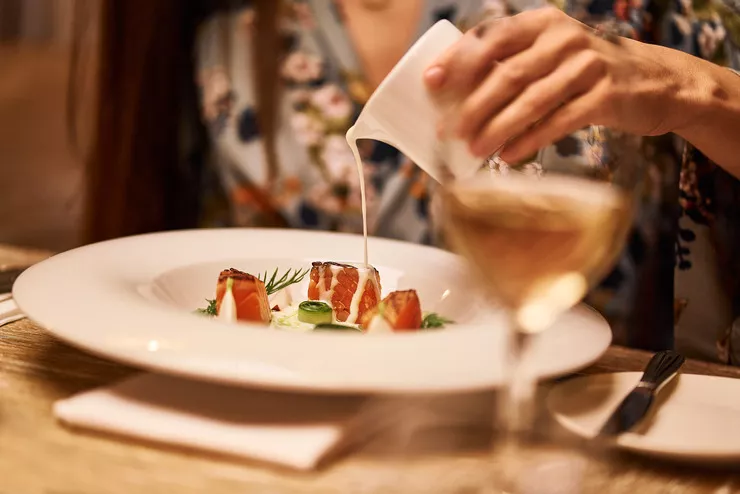 A view of a white plate with three pieces of fish on an elegantly set table. The dish is garnished with green sprigs and some vegetables. The woman sitting behind the table is rising her hand with a pitcher and pouring some sauce over her fish. A blurred glass of white wine is standing on the right in the foreground.