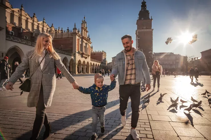 A young couple with a small child are walking briskly in the Main Market Square in Kraków. There is a flock of pigeons on the square's floor behind them on the right, the Town Hall Tower peers from behind the man’s shoulder, while the Cloth Hall provides the background for the woman. It is a sunny day, and the setting sun is casting long shadows.