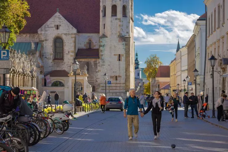Blick auf eine Straße in der Altstadt mit einem älteren Ehepaar im Vordergrund. An der Seite sind stehende Fahrräder, Skulpturen an der Kirchenmauer und eine Reihe von Mietshäusern entlang der Straße zu sehen. Die St. Andreaskirche ist im Hintergrund zu sehen.