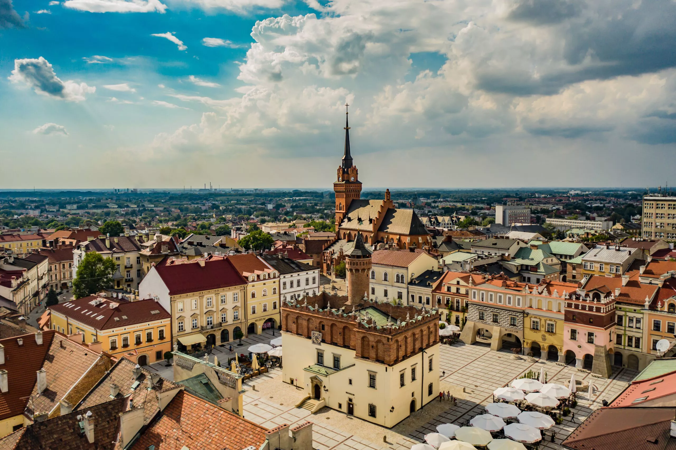 An aerial view of the Tarnów market square. Tarnów town hall stands In the centre of the square, surrounded by townhouses. The whole is dominated by the tall spire of the cathedral soaring from among more townhouses into the white clouds that are visible in the blue sky.
