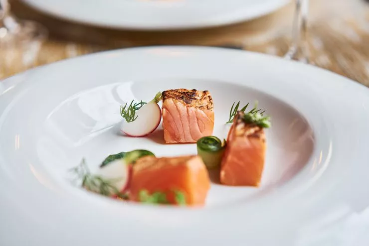 Blick auf einen weißen Teller mit drei quadratischen Fischstücken. Das Gericht ist mit grünen Gewürzzweigen und Gemüsestücken dekoriert.