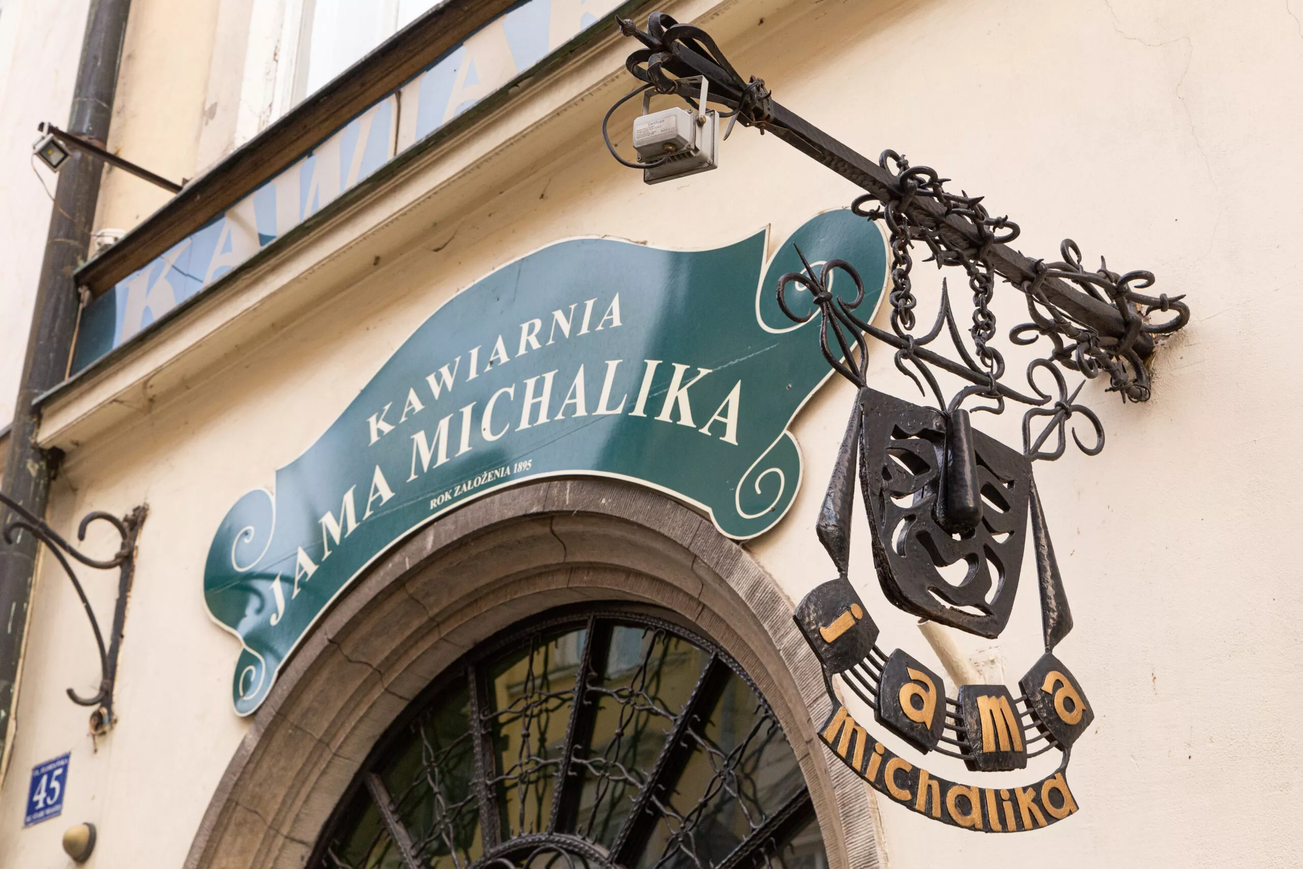 A small section of a townhouse façade with a fancy green sign reading “Kawiarnia Jama Michalika” fixed above the top of a gate, next to an elaborated metal café sign hanging from a rod.