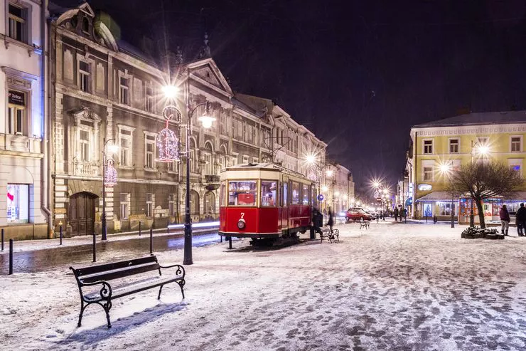 A night view of a snowy city street with a bench, lamps, and some distant trees. Standing centrally is a red, vintage tramcar without wheels. The houses under the black sky providing the background are basked in artificial lighting.