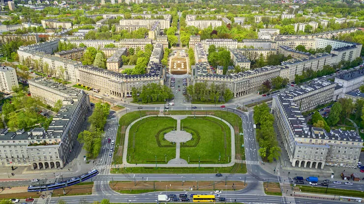 A distant drone-eye view of a square surrounded by buildings, with streets radiating symmetrically from it. The centre of the square is taken by a lawn intersected with paths. There are trees growing inside the city blocks, and the streets are busy with cars and trams.