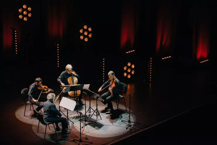 Luftaufnahme einer Gruppe von Musikern, die auf einer verdunkelten Bühne sitzen und Saiteninstrumente spielen. Vor den Musikern stehen Pulte mit Notenblättern.