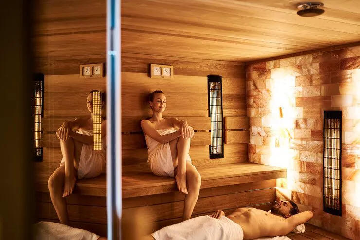 A look into a sauna illuminated with soft light. A woman wrapped in a towel is sitting on a wooden bench, while a man wrapped in another towel is lying at her feet. Both have closed eyes.