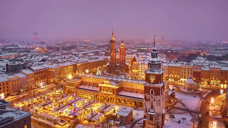 A drone-eye view of Kraków's Main Market Square at dusk on a winter evening. The brightly illuminated Christmas market stalls take the bottom left-hand corner are surrounded by a line of townhouses, the Cloth Hall, and the Town Hall Tower, with the façade of St Mary's Church peering from behind the centrally photographed Cloth Hall. Everything is gently sprinkled with snow.