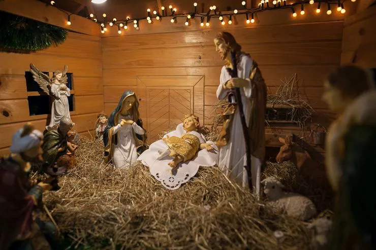 A peek into the interior of a wooden nativity scene with figures of Blessed Virgin, St Joseph, and Infant Jesus lying centrally on a doily covering straw. They are surrounded by figures of angels, shepherds, and farm animals standing on the straw by the walls of the cottage.