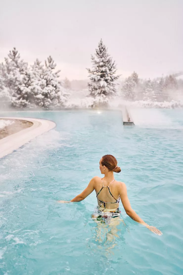 A view of a hot pool surrounded by snow-covered trees. The centre of this vertical photo is taken by a woman in a swimsuit, turned to us with her back, standing in the pool.