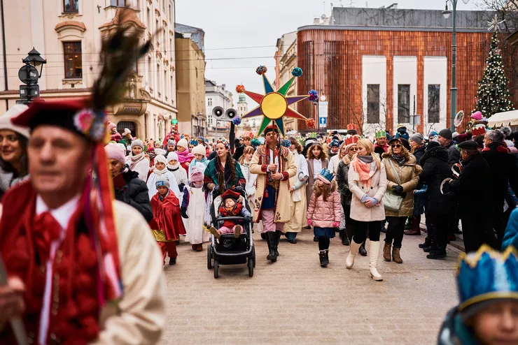 A colourful procession is going up Grodzka street. The person in the centre is walking among a crowd of people and carrying a large colourful star on a tall pole. There are some buildings on the left, and Wyspiański Pavilion and a huge Christmas tree on the right.